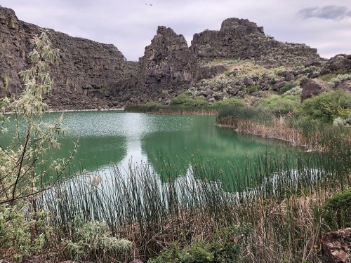 A serene green lake surrounded by rocky cliffs and lush vegetation under a cloudy sky.