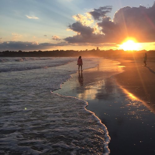 A person walks along a beach at sunset, with waves gently lapping at their feet and clouds in the sky.