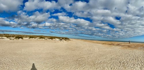 A wide sandy beach under a blue sky filled with fluffy clouds, with a distant figure walking along the shore.
