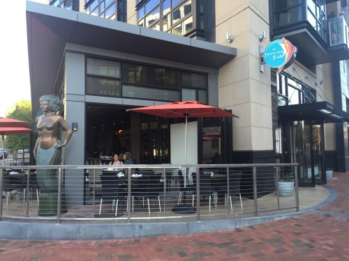 A modern restaurant exterior with outdoor seating, featuring a large statue of a woman and red umbrellas.