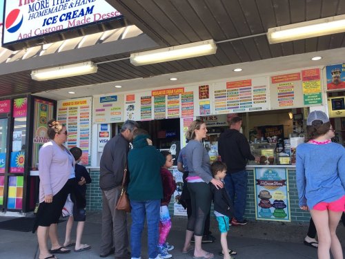 A busy ice cream shop with a line of customers waiting to order, colorful menu signs displayed above the counter.
