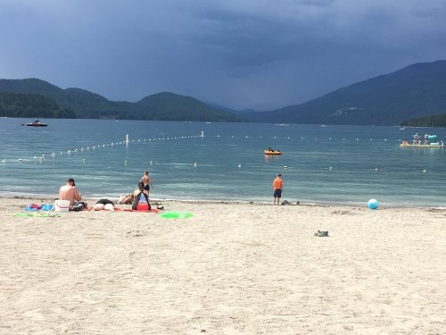 A sandy beach with people relaxing, swimming, and a stormy sky in the background over a calm lake.