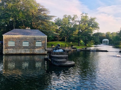A serene park scene featuring a stone building, a statue on a fountain, and a gazebo across a calm pond.