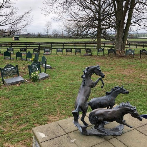 A sculpture of three rearing horses in a grassy area with gravestones in the background under a cloudy sky.