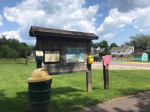 A wooden information board with maps and a phone, next to a trash can, in a grassy area under a cloudy sky.