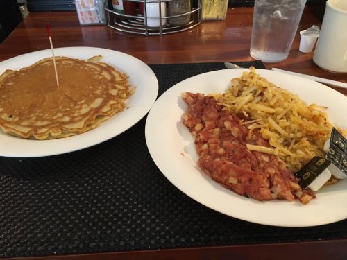 A plate with a large pancake and another plate with hash browns and a meat dish, served at a diner.