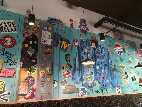 Colorful wall covered in retro stickers, patches, and memorabilia, featuring a large denim jacket and playful decor.