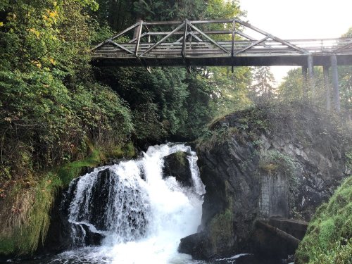 A wooden bridge spans over a waterfall surrounded by lush greenery and rocky terrain.