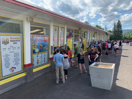 A long line of people waiting outside a shaved ice stand on a sunny day, with a menu displayed on the wall.