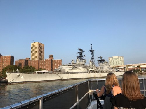 Two people on a boat view a large gray battleship docked near city buildings under a clear blue sky.