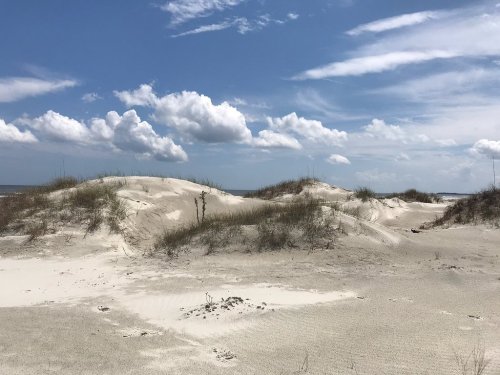 A sandy beach landscape with dunes, sparse grass, and a blue sky dotted with fluffy white clouds.