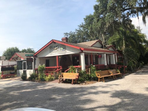 A charming, red-accented building with a sign, surrounded by greenery and benches, set in a gravel area.
