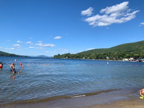 A serene lake scene with people swimming, surrounded by green hills and a clear blue sky dotted with clouds.