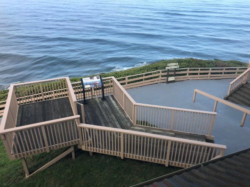 A wooden observation deck with railings overlooking a calm ocean, surrounded by greenery and a clear sky.