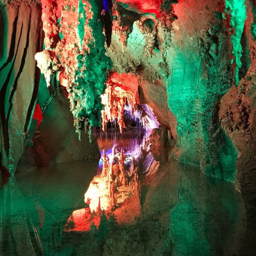 A colorful cave with stalactites and stalagmites reflected in still water, illuminated by green and red lights.