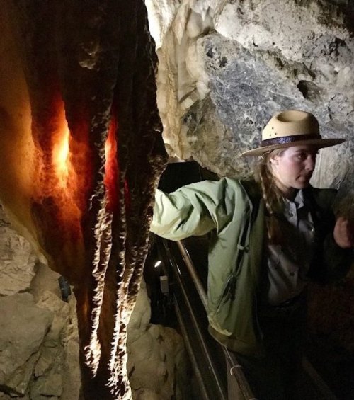 A park ranger stands near a glowing rock formation in a cave, illuminated by soft light.