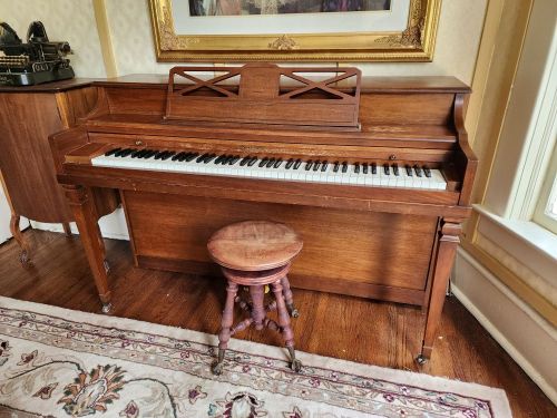 A wooden piano with a matching stool, set in a room with elegant decor and a framed artwork in the background.