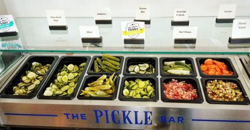 A variety of pickled vegetables displayed in containers at a pickle bar, including cucumbers, okra, and carrots.