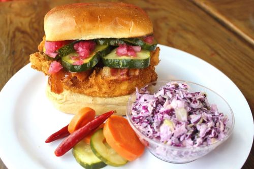 A crispy fried chicken sandwich topped with pickles and onions, served with coleslaw and pickled vegetables on the side.