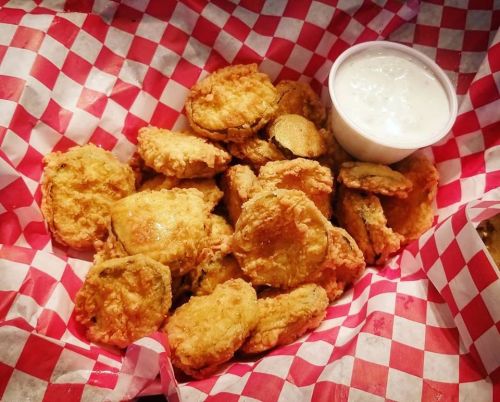A basket of crispy fried pickles with a side of dipping sauce, lined with red and white checkered paper.