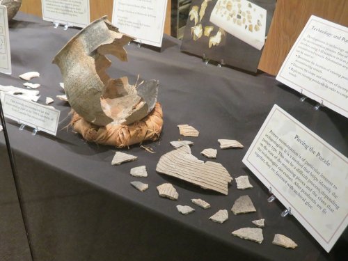 Display of ancient pottery shards and a reconstructed pot on a table, with informational signs in the background.