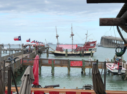 A wooden pirate ship docked at a pier with flags, surrounded by water and a cloudy sky.