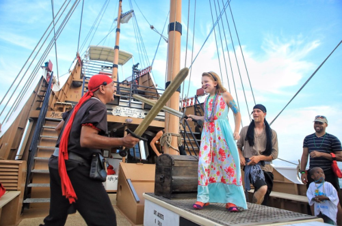 A child in a colorful dress playfully duels with a pirate on a ship, while other adults watch and smile.