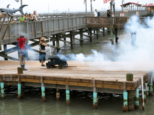 Two performers in pirate costumes fire a small cannon on a wooden pier, with smoke billowing into the air.