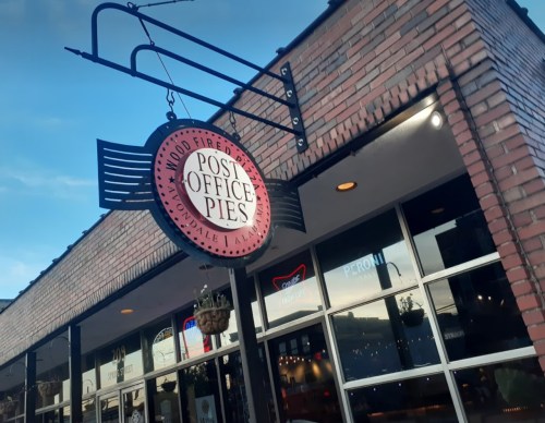 Sign for "Post Office Pies" hanging above a brick building with large windows, under a blue sky.