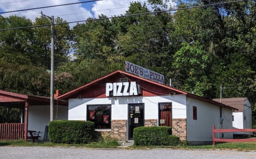 A small pizza restaurant with a red roof and sign, surrounded by greenery and gravel parking.