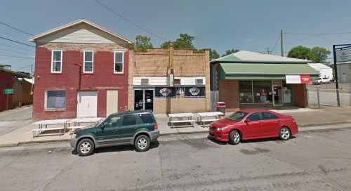A street view of a brick building with a green roof, parked cars, and outdoor seating along the sidewalk.