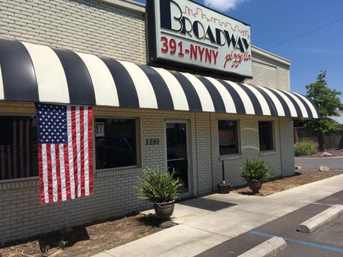 Exterior of Broadway Pizzeria with a striped awning and an American flag hanging by the entrance on a sunny day.