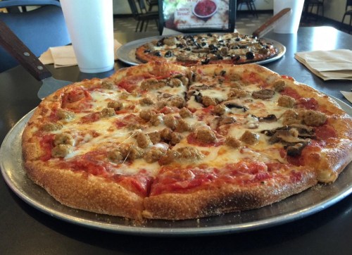 Two pizzas on metal trays: one topped with sausage and cheese, the other with olives and mushrooms, on a restaurant table.