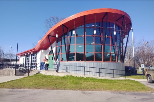 Modern building with a curved red roof, large windows, and two people standing outside on a sunny day.