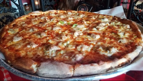 A large pizza with melted cheese, sausage, and green peppers on a silver tray, set on a checkered tablecloth.