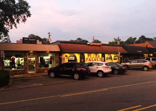 A street view of a pizza restaurant and a formal wear shop, with parked cars and a twilight sky.