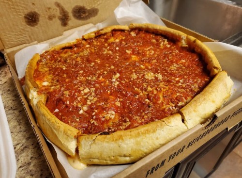 A deep-dish pizza in a cardboard box, topped with a thick layer of tomato sauce and sprinkled with cheese and herbs.