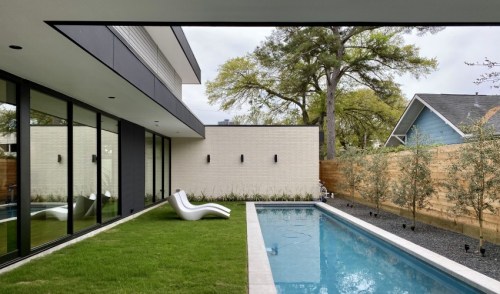 Modern backyard with a pool, sleek lounge chairs, grass, and landscaped greenery, framed by large glass windows.