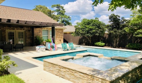 A backyard with a pool, spa, lounge chairs, and colorful pool floats, surrounded by greenery and a brick house.