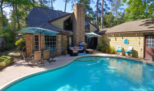 A sunny backyard with a pool, patio furniture, and umbrellas near a cozy house surrounded by trees.