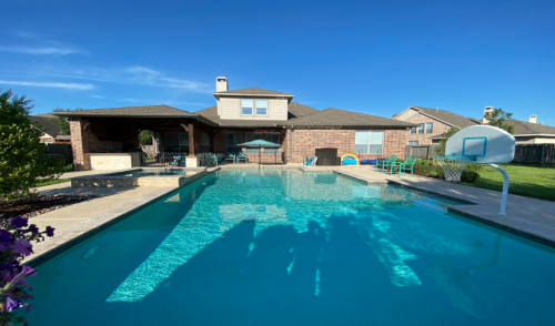 A clear blue pool in a backyard with a brick house, patio furniture, and a basketball hoop under a sunny sky.