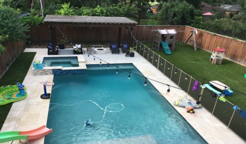 Aerial view of a backyard pool area with a hot tub, playset, and green lawn, surrounded by string lights and safety fencing.