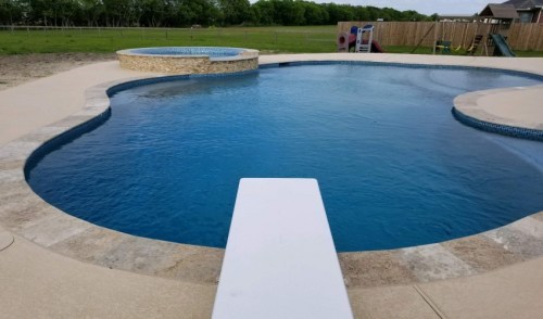 A clear blue swimming pool with a diving board, surrounded by a patio and a grassy area in the background.