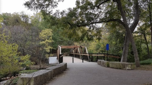 A wooden bridge spans a path in a park, surrounded by trees with autumn foliage.