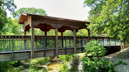 A wooden covered bridge spans a small creek, surrounded by lush greenery and trees.