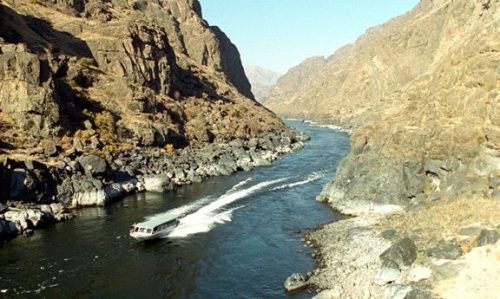 A boat navigates a winding river surrounded by steep, rocky mountains under a clear blue sky.