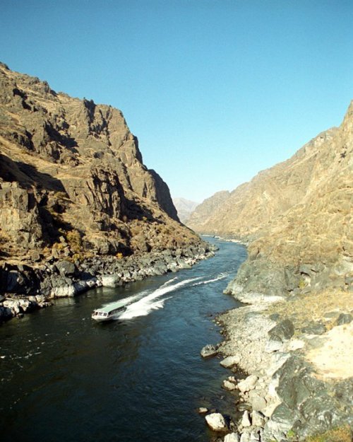 A boat navigates a winding river surrounded by steep, rocky mountains under a clear blue sky.