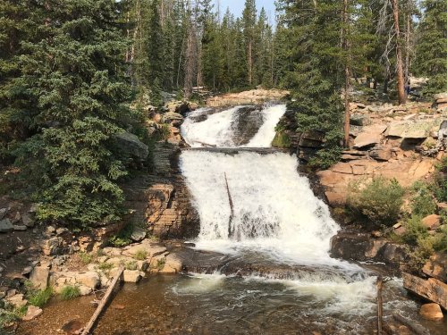 A cascading waterfall flows over rocky terrain, surrounded by lush green trees and a serene natural landscape.