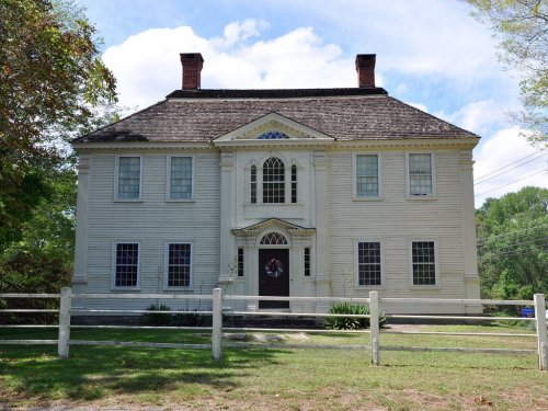 A large, historic white house with a symmetrical facade, featuring a central door and decorative windows, surrounded by greenery.