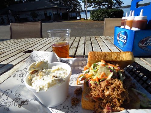 A plate with pulled pork sandwich, coleslaw, and a side of potato salad, with a drink on a wooden table by a lake.